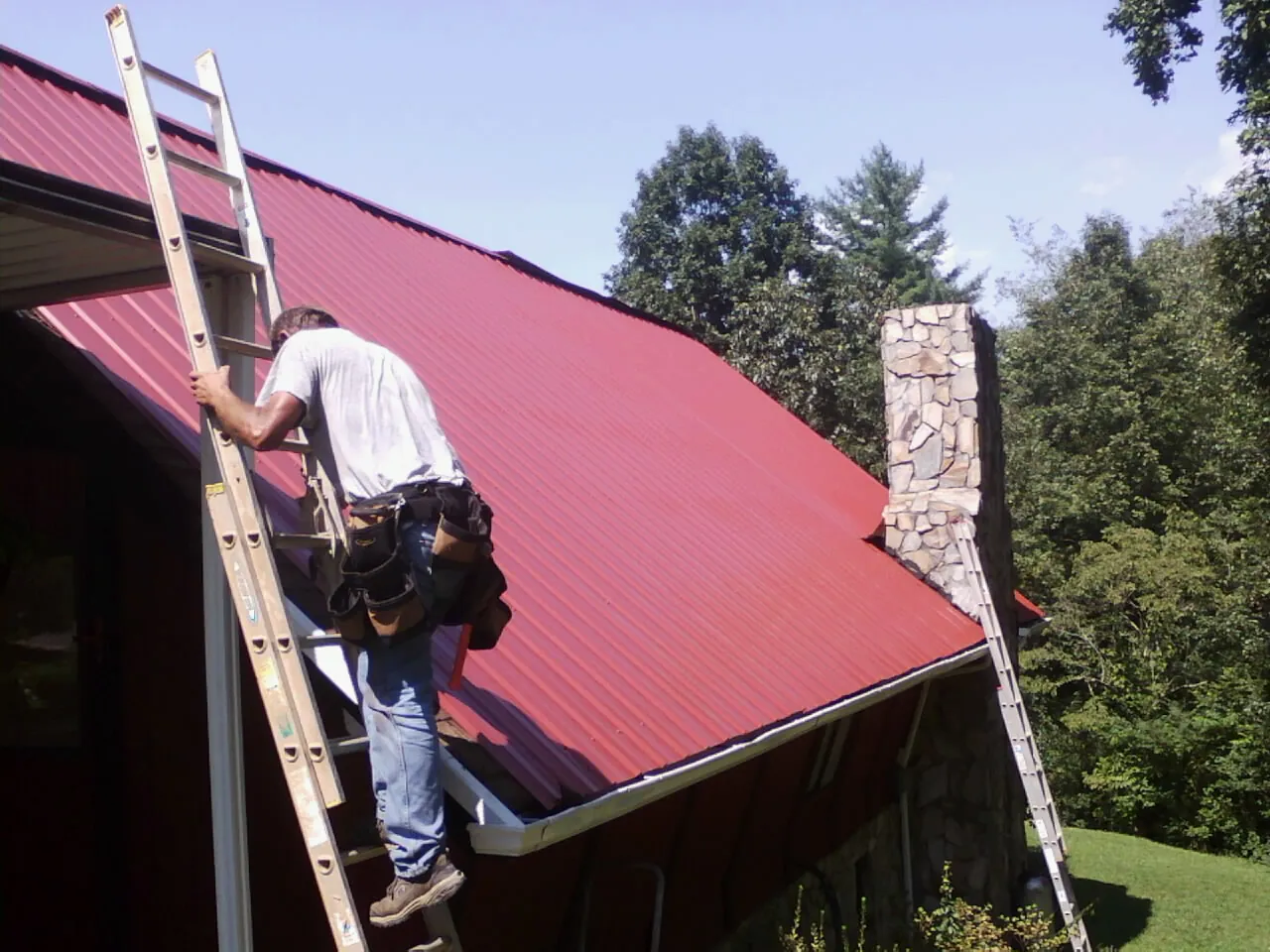 Roofer on a ladder beside a red metal roof and stone chimney — Soffit & Fascia Repair by Roofing Brothers in Pruden